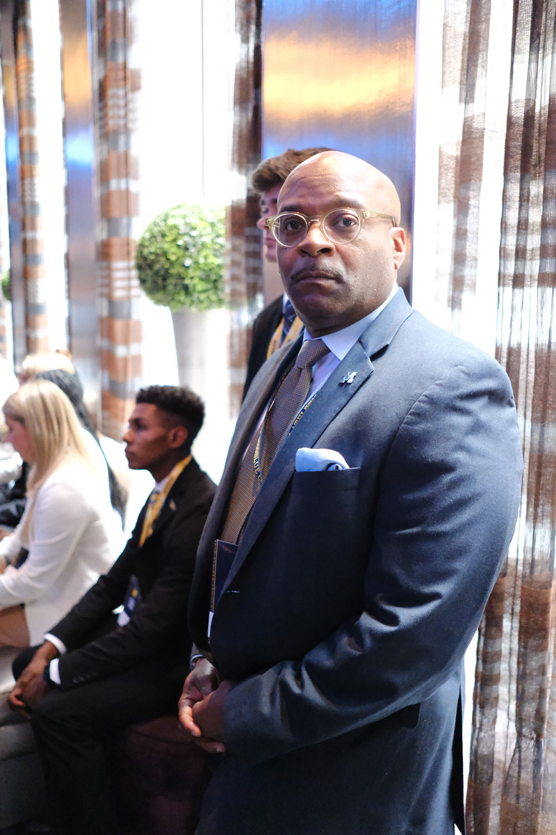 A man in a blue suit and glasses seated beside a bright window