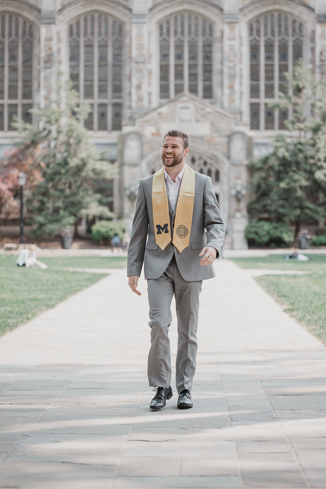 A graduate in a gray suit walking along a campus path